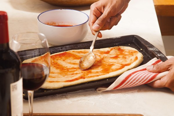 Mão masculina espalhando molho de tomate com uma colher sobre a massa de pizza caseira em uma forma preta. Ao lado, um bowl branco com molho e uma taça de vinho tinto.